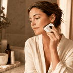Woman in a bathrobe using a Gua Sha skincare device in a bathroom setting