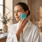 Woman in a white robe using a blue facial cleansing brush in a bathroom.