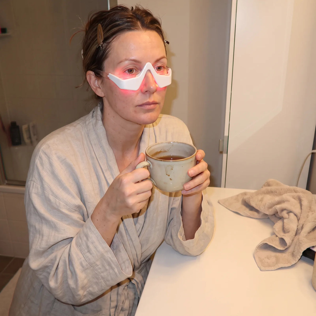A woman sits at her bathroom counter with a mug of tea, wearing the Halo under-eye device as it glows red.