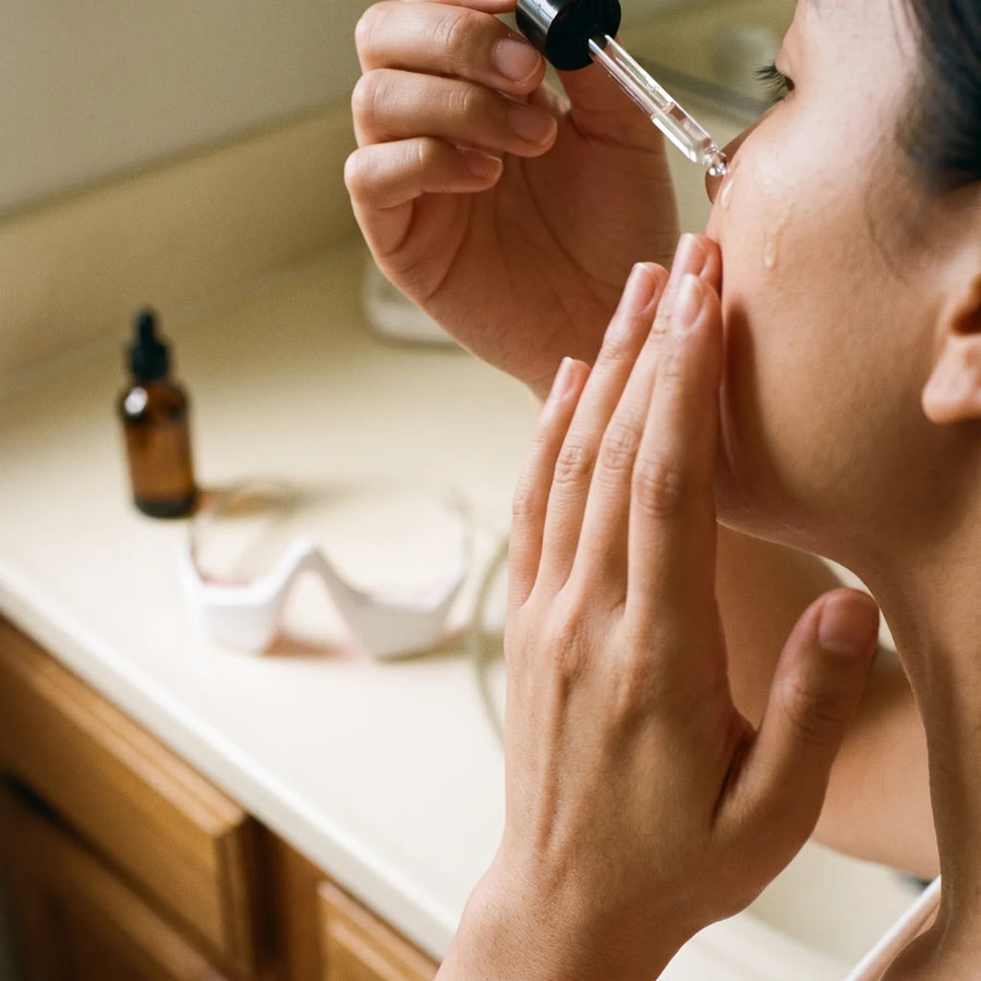 A woman at her bathroom counter, leaning toward the mirror as she works a few drops into her cheek.